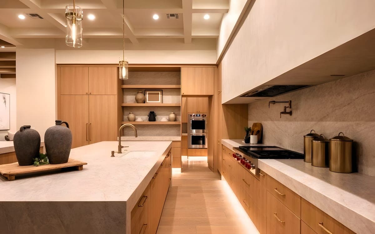 Kitchen from the working side showing a large island with undermount sink, double wall ovens, gas range with pot filler, white oak cabinetry to ceiling, and brass hardware