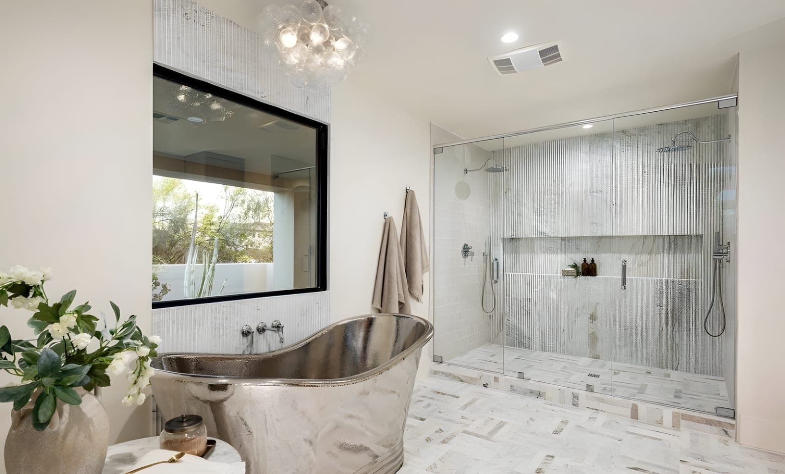 Freestanding polished nickel soaking tub centered beneath a glass bubble chandelier, with a fluted marble shower wall and frameless glass enclosure beyond