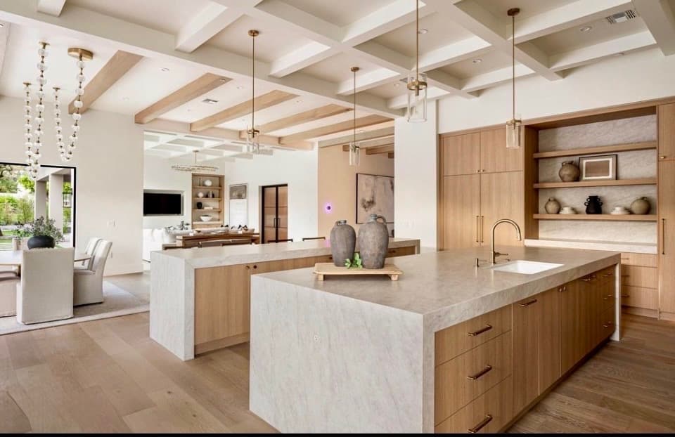 Open-plan kitchen and great room with dual white oak islands, honed stone countertops, brass hardware, globe pendants, and coffered ceiling with exposed wood beams