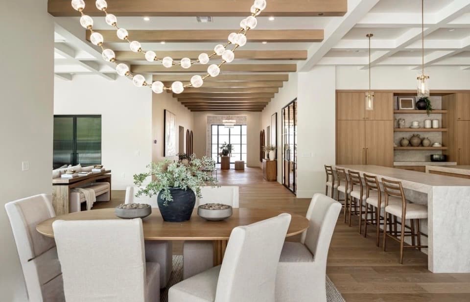 Dining area with an oval white oak table, upholstered chairs, and a sweeping globe pendant light installation across a coffered ceiling, adjacent to the kitchen