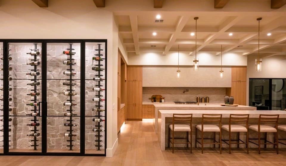 Kitchen with illuminated iron-framed wine storage wall, stone backing, white oak cabinetry, and honed stone island under a coffered beam ceiling