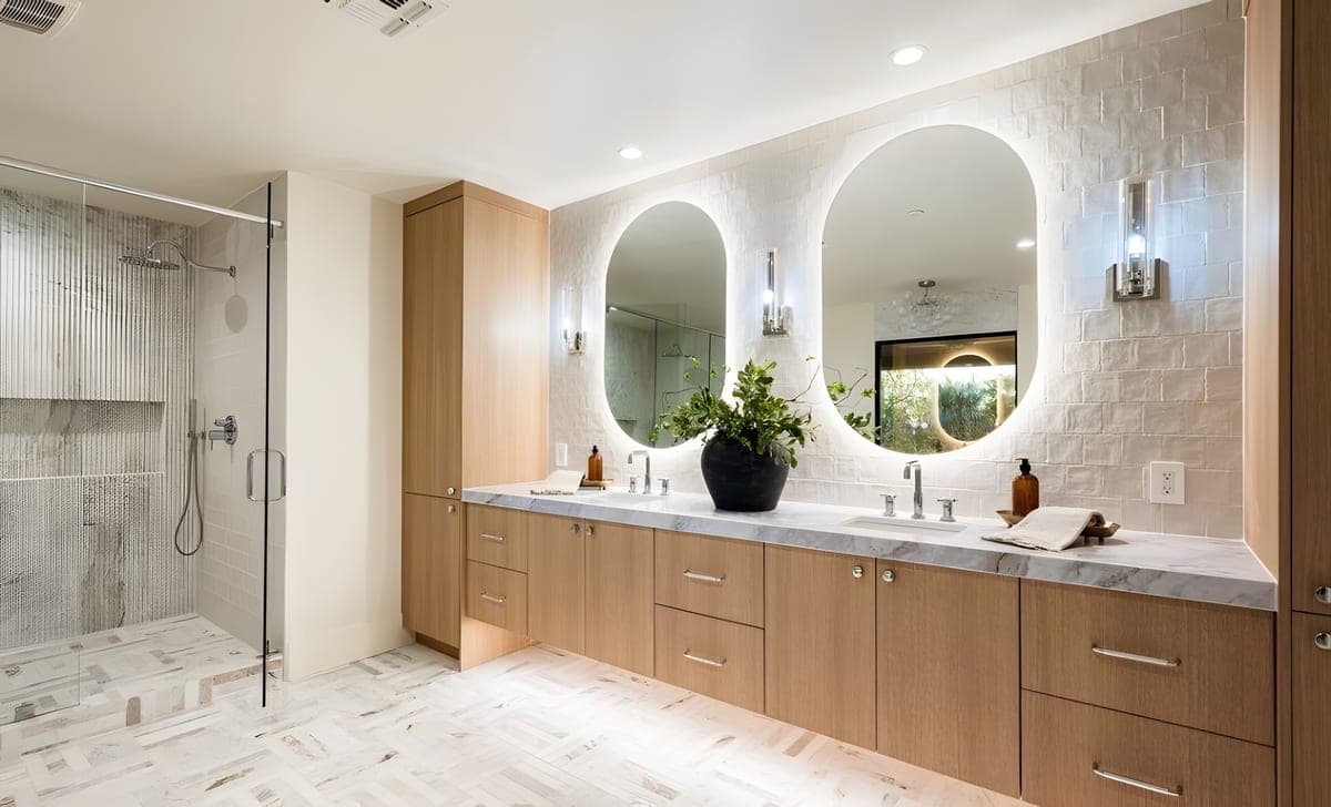 Double vanity in white oak with dual backlit oval mirrors, zellige tile walls, and a fluted marble shower visible in the background