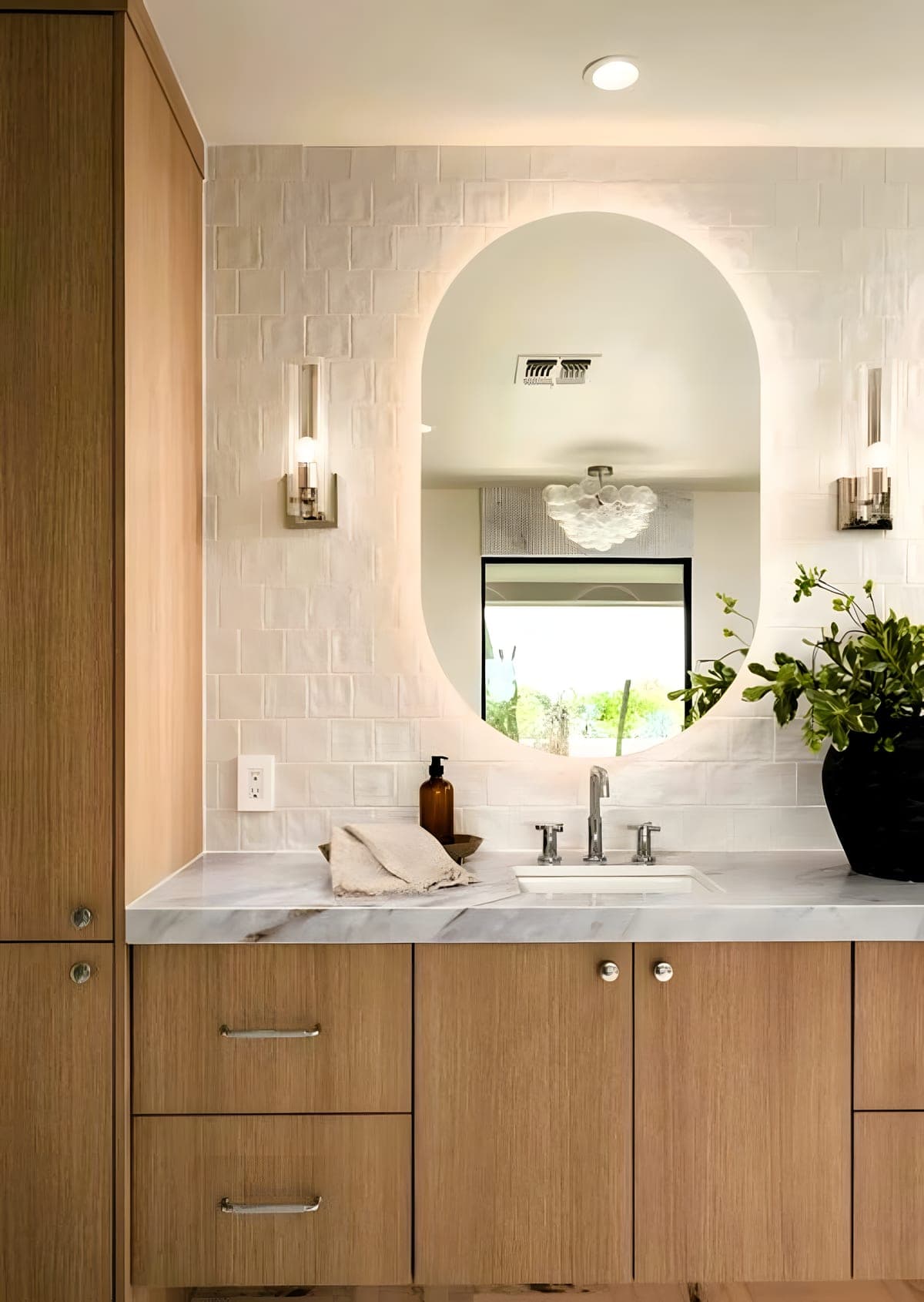 Single vanity with white oak cabinet, honed marble countertop, and a backlit oval mirror against a zellige tile wall