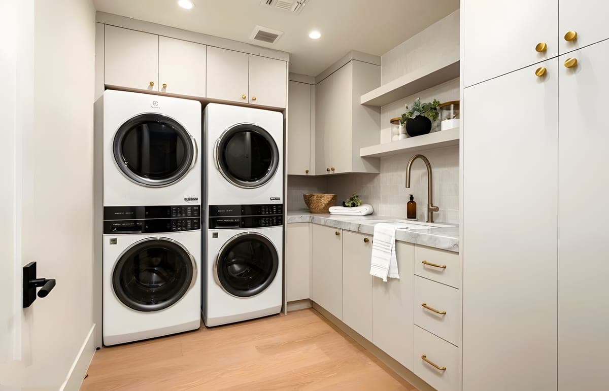 Laundry room with dual stacked washer-dryer units, white cabinetry with brass hardware, marble countertop, and a utility sink with brass faucet